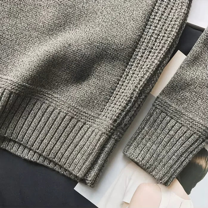 Pull en laine gris texturé avec manches longues, détails côtelés, posé sur des magazines. Mode automne-hiver, vêtement chaud et élégant.