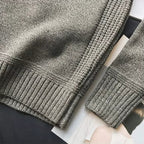 Pull en laine gris texturé avec manches longues, détails côtelés, posé sur des magazines. Mode automne-hiver, vêtement chaud et élégant.