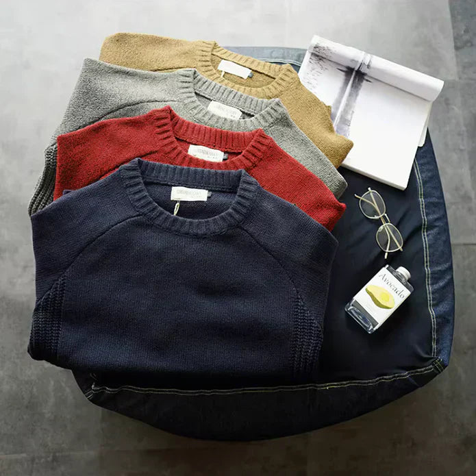 Quatre pulls en laine de couleurs différentes (bleu, rouge, gris, beige) posés sur un pouf, avec lunettes, livre et paquet de cigarettes à côté.