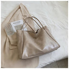 Sac à main en cuir beige posé sur un manteau crème, avec un livre "Matisse" et un verre en cristal, sur un lit blanc. Accessoire mode élégant.