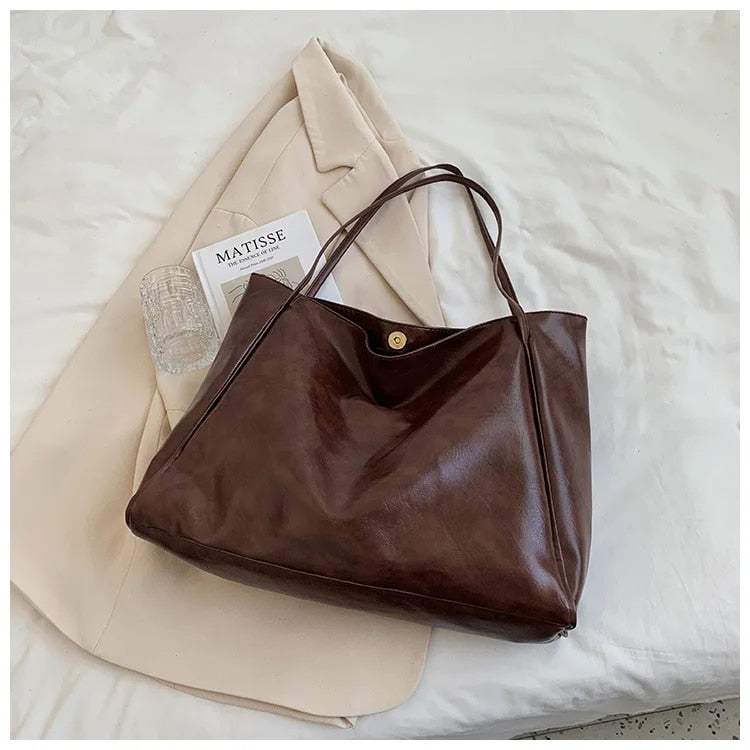 Sac à main en cuir marron sur un manteau beige, posé sur un lit blanc. Accessoires élégants, style chic, mode féminine, tendance automne.