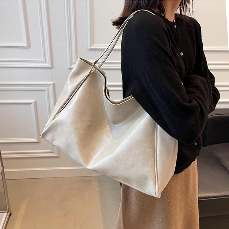 Sac à main en cuir beige porté par une femme en pull noir et jupe marron, devant un mur blanc élégant. Accessoire mode chic et tendance.