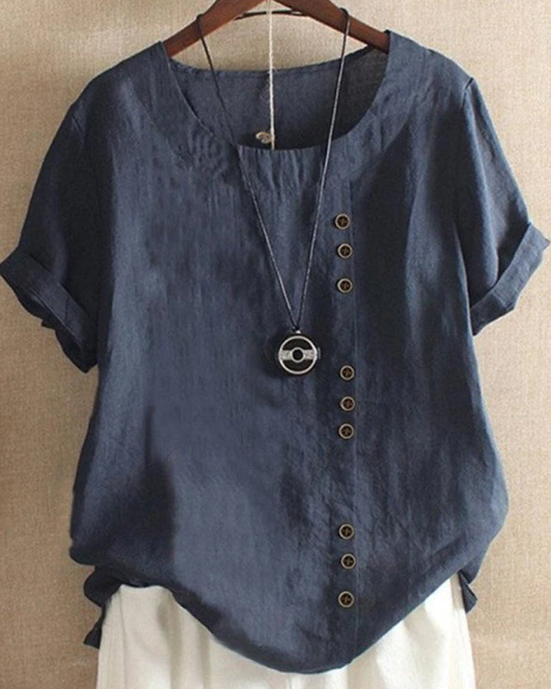 Blouse en lin bleu avec boutons décoratifs asymétriques, manches courtes retroussées, col rond, suspendue sur un cintre. Mode féminine élégante.