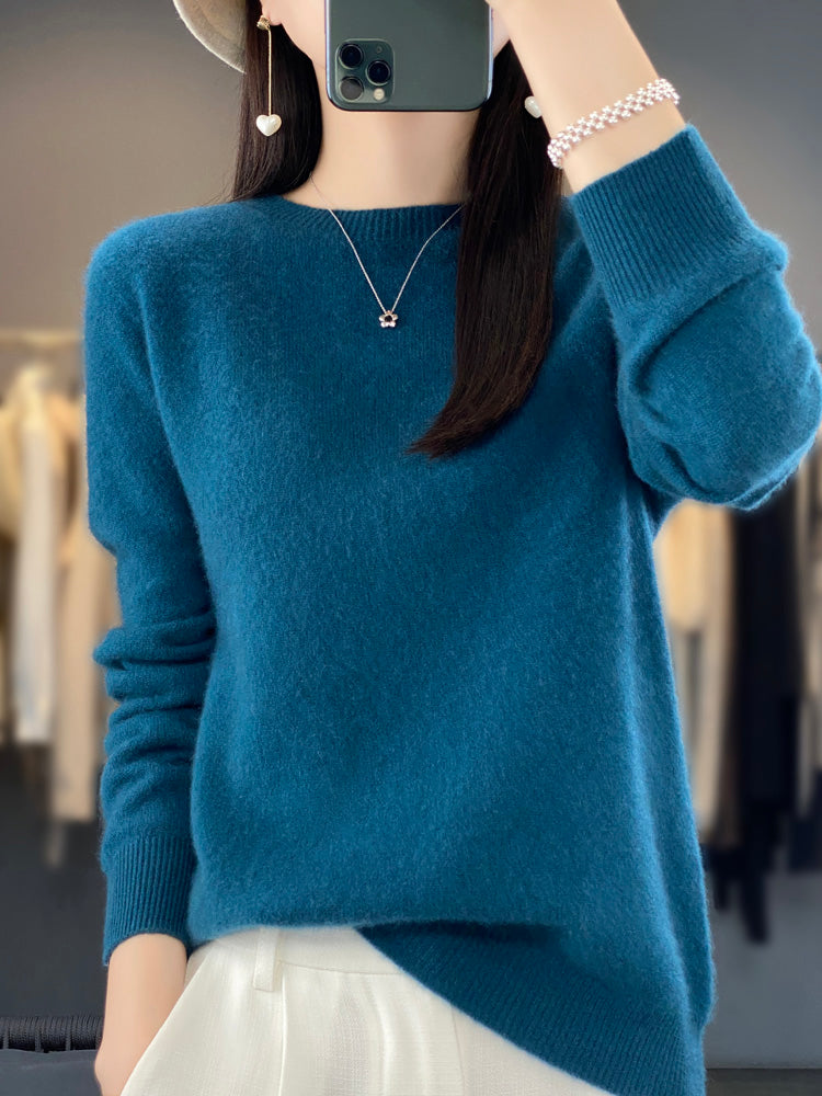 Femme en pull bleu en laine, tenant un smartphone, portant un collier étoile et un bracelet perlé, mode automne-hiver, style décontracté.