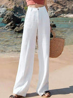 Femme en pantalon blanc ample sur plage, tenant un sac en osier. Mode estivale, tenue décontractée, bord de mer, style chic et tendance.