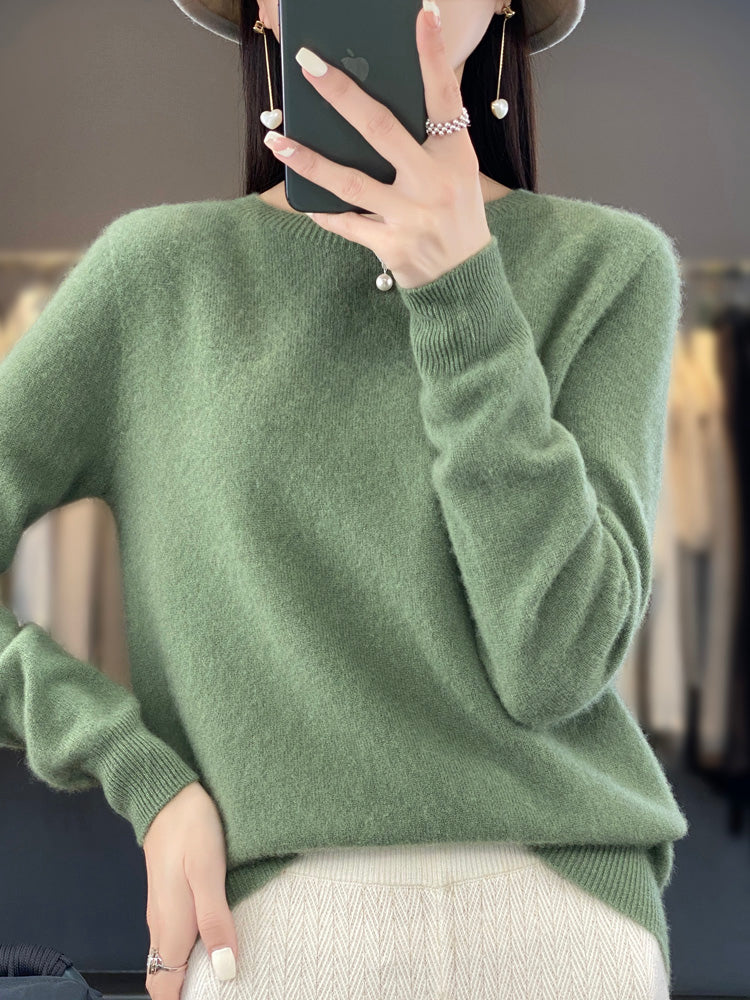 Femme portant un pull vert en cachemire, tenant un smartphone, avec un chapeau et des boucles d'oreilles perle. Mode automne-hiver élégante.