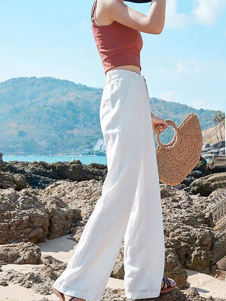 Femme en pantalon blanc ample et top rouge, tenant un sac en paille, sur une plage rocheuse ensoleillée avec vue sur la mer et les collines.