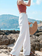 Femme en pantalon blanc ample et top rouge, tenant un sac en paille, sur une plage rocheuse ensoleillée avec vue sur la mer et les collines.