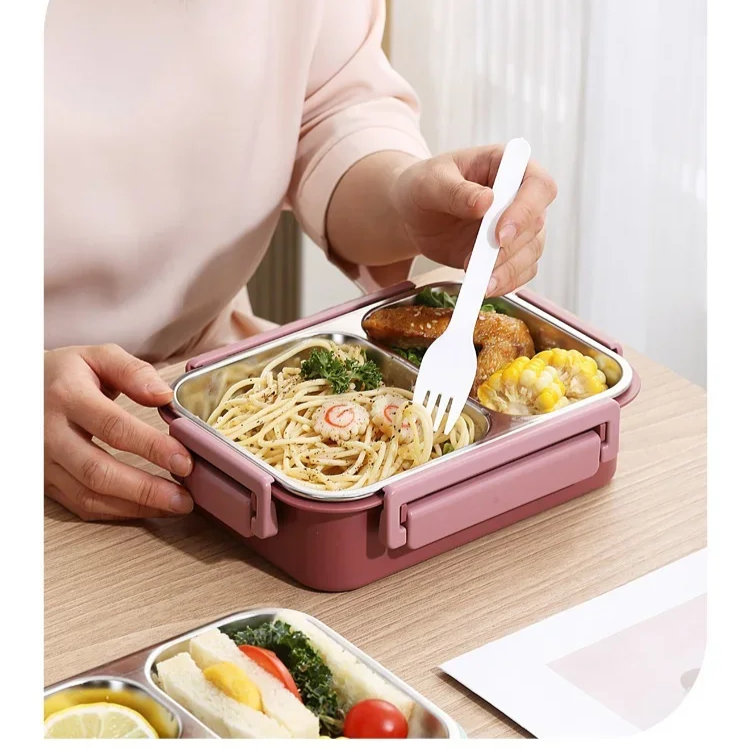 Personne mangeant un déjeuner dans une boîte bento rose avec des compartiments contenant des pâtes, du poulet, du maïs et des légumes. Repas équilibré.