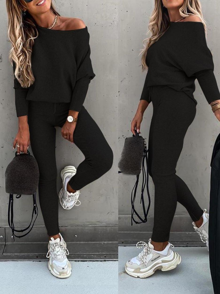 Femme en ensemble noir décontracté, pull ample et pantalon ajusté, portant des baskets blanches et un sac à main pelucheux, posant contre un mur gris.