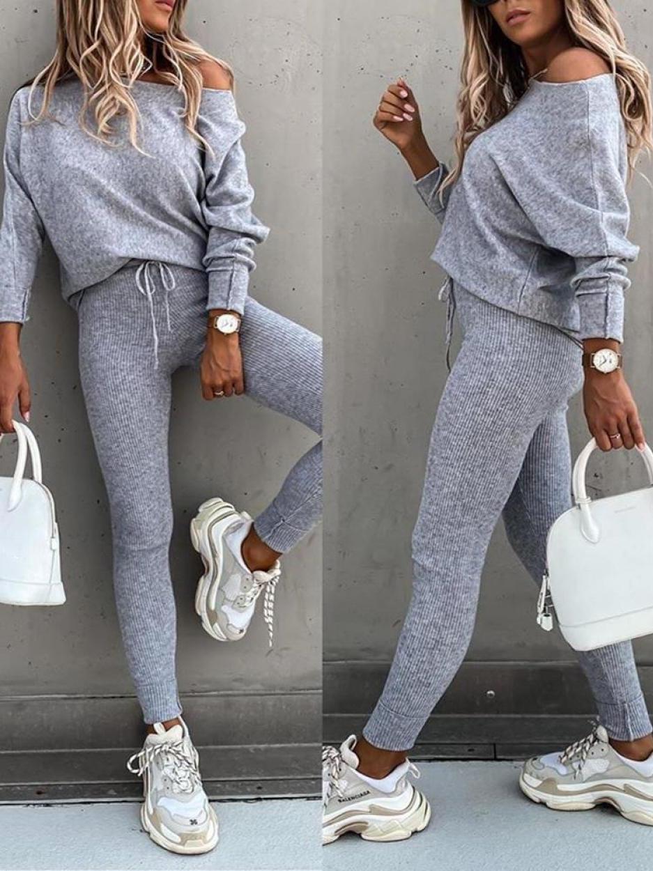 Femme en tenue décontractée grise, pull ample et leggings, portant des baskets blanches et un sac à main blanc, posant contre un mur gris.