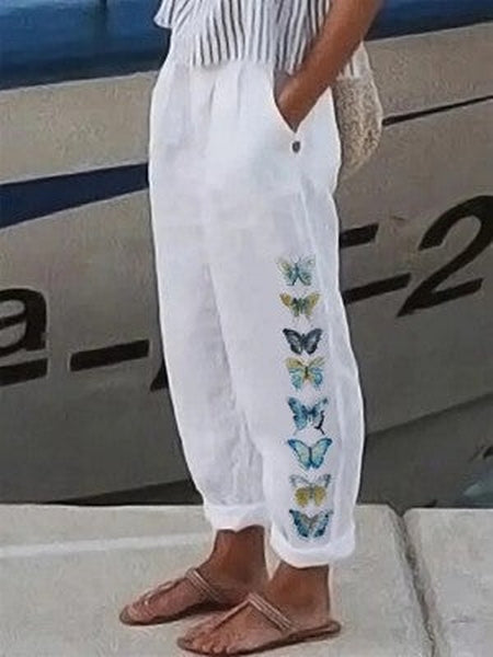 Pantalon blanc avec motifs de papillons colorés sur le côté, porté par une personne debout près d'un bateau. Mode estivale, style décontracté.