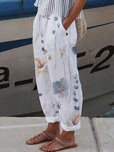 Pantalon blanc à motifs floraux, femme debout sur un quai, sandales marron, style décontracté estival, fond de bateau flou.