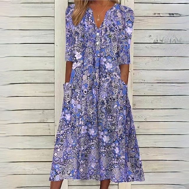 Robe longue fleurie bleue et violette avec manches trois-quarts, poches latérales, sur fond de bois blanc. Mode féminine élégante et décontractée.