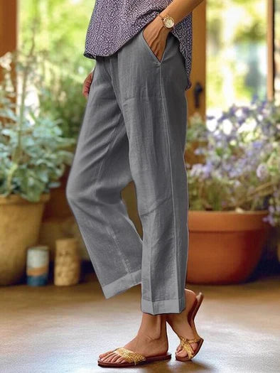 Pantalon en lin gris pour femme, style décontracté, poches latérales, porté avec des sandales dorées, ambiance estivale, fond de plantes vertes.