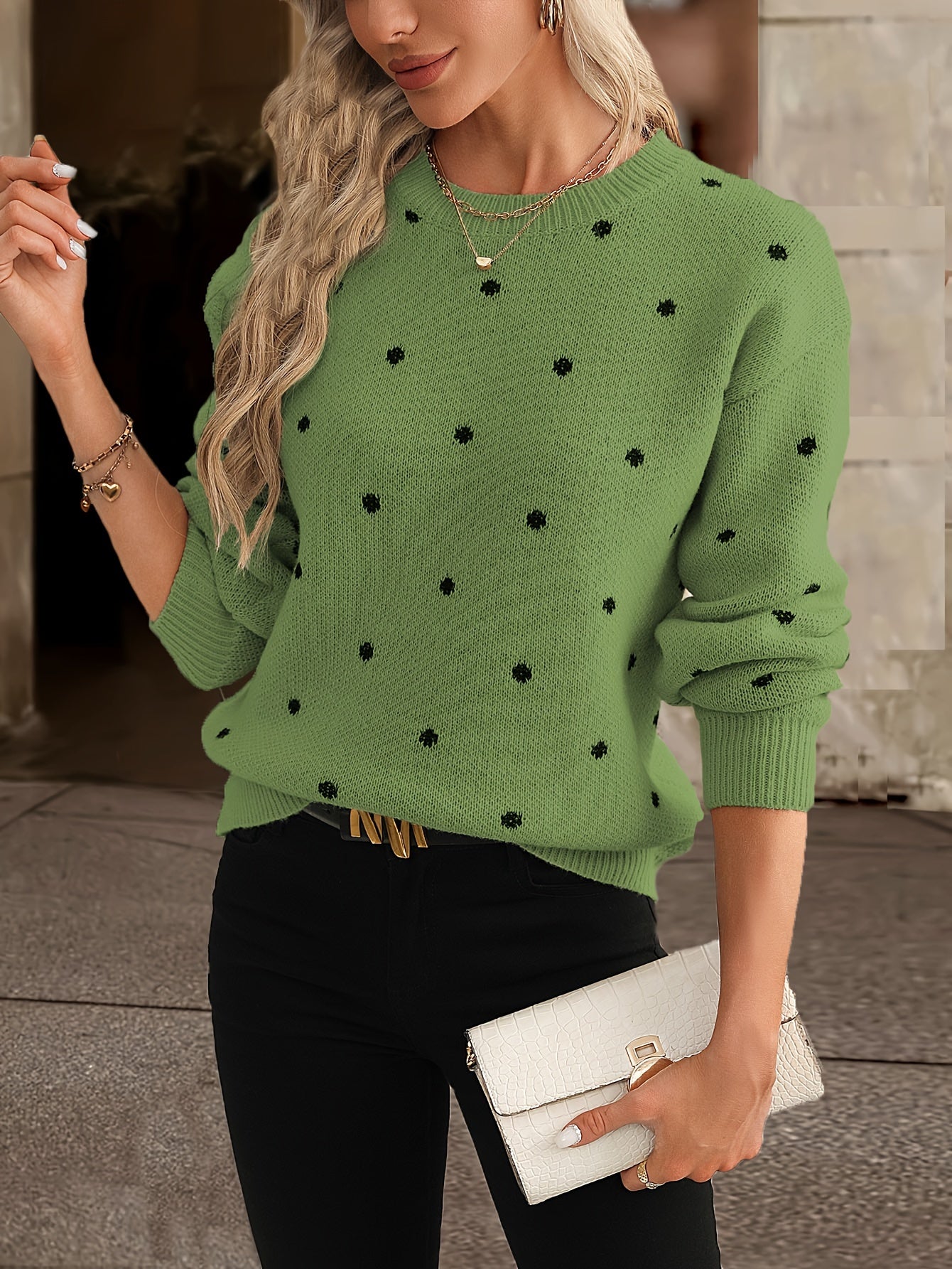Femme portant un pull vert à pois noirs, tenant une pochette blanche. Mode automne, style décontracté chic, accessoires dorés.
