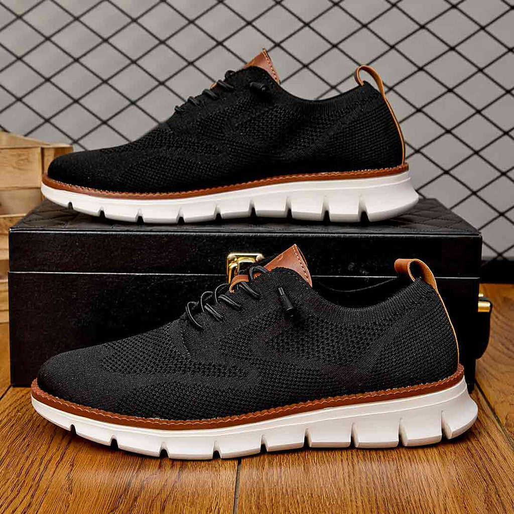 Chaussures noires en maille respirante avec semelle blanche, posées sur une boîte noire en bois, sur un sol en bois, fond à motif géométrique.