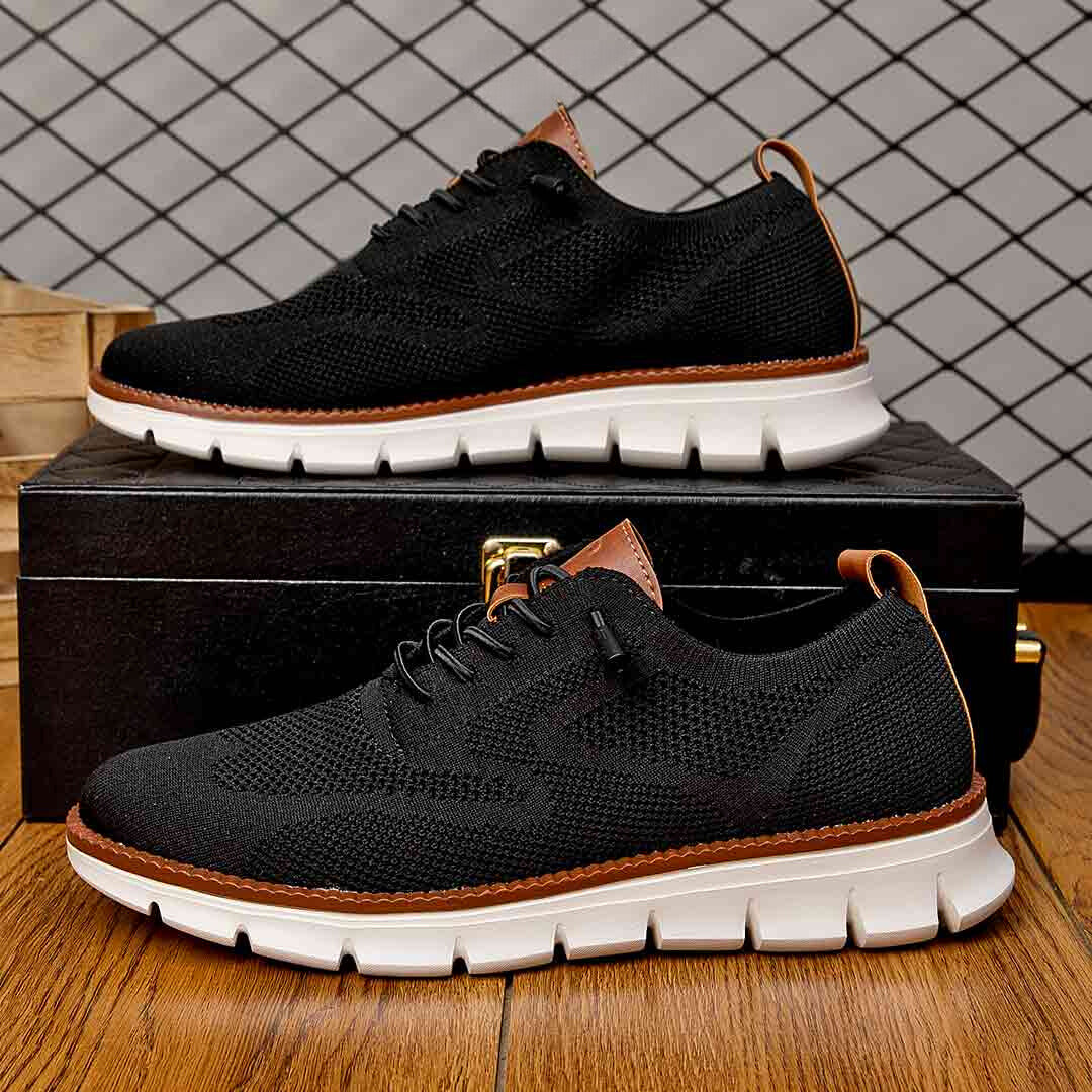 Chaussures noires en maille respirante avec semelle blanche, posées sur une boîte noire en bois, sur un sol en bois, fond à motif géométrique.