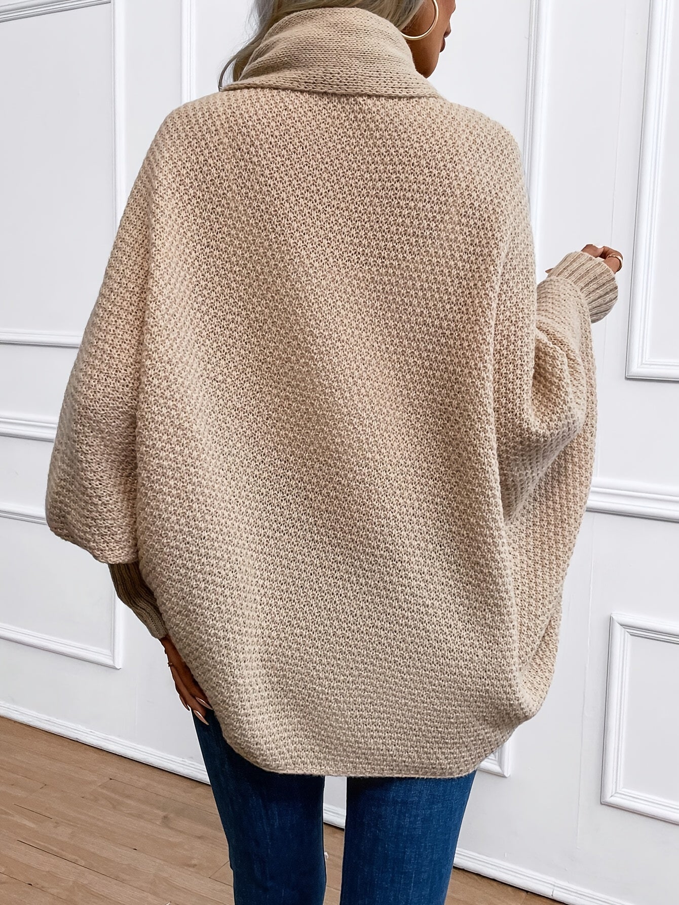 Femme portant un pull oversize beige en tricot texturé, col roulé, manches longues, devant un mur blanc. Mode automne-hiver, style décontracté.