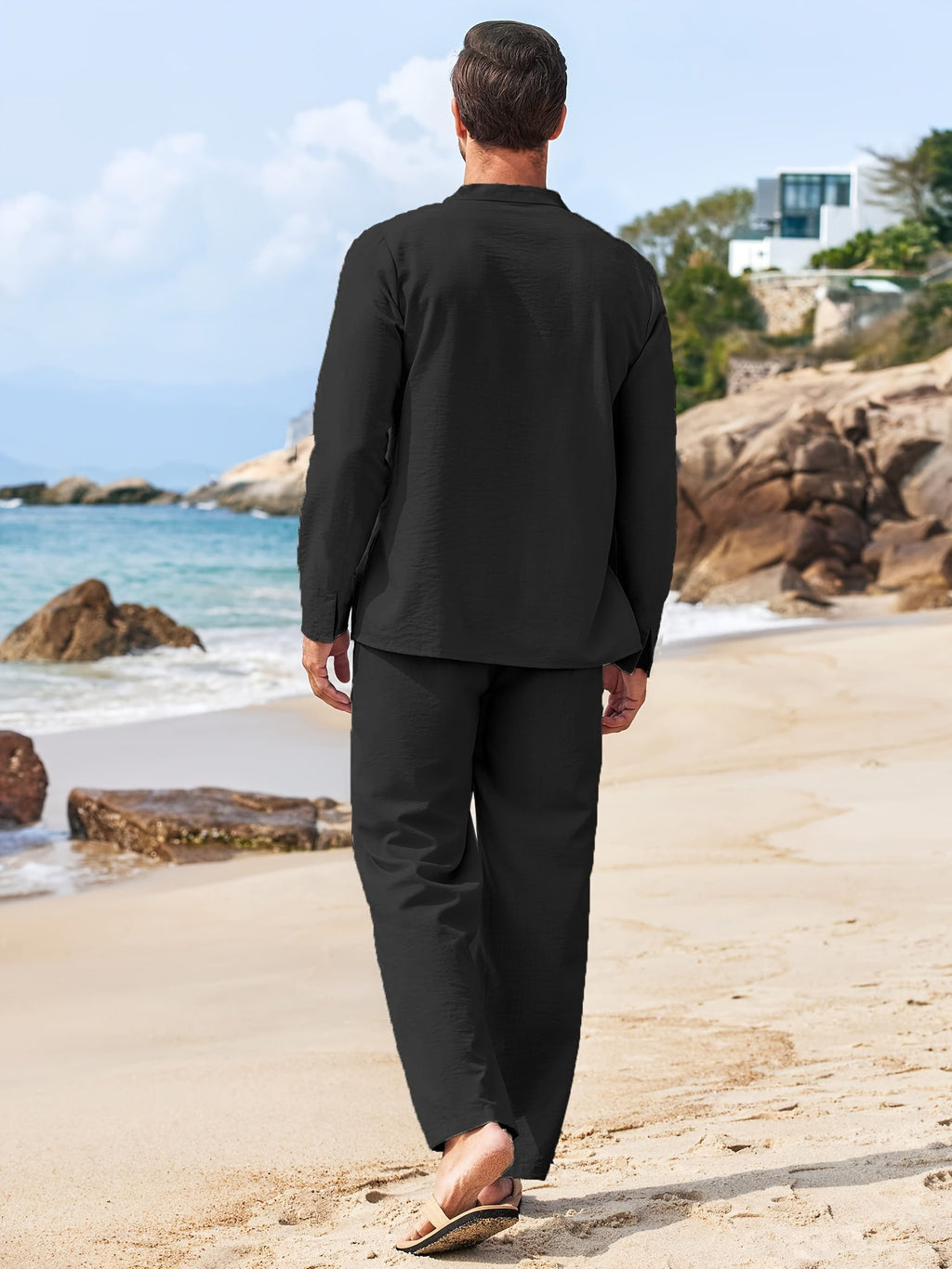 Homme en tenue noire décontractée marchant sur une plage, vue de dos, ciel bleu et maisons en arrière-plan. Mode masculine, vacances, bord de mer.