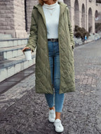 Femme portant un manteau long vert matelassé, jean slim bleu, pull blanc, baskets blanches, tenant un gobelet à café, marchant en ville.
