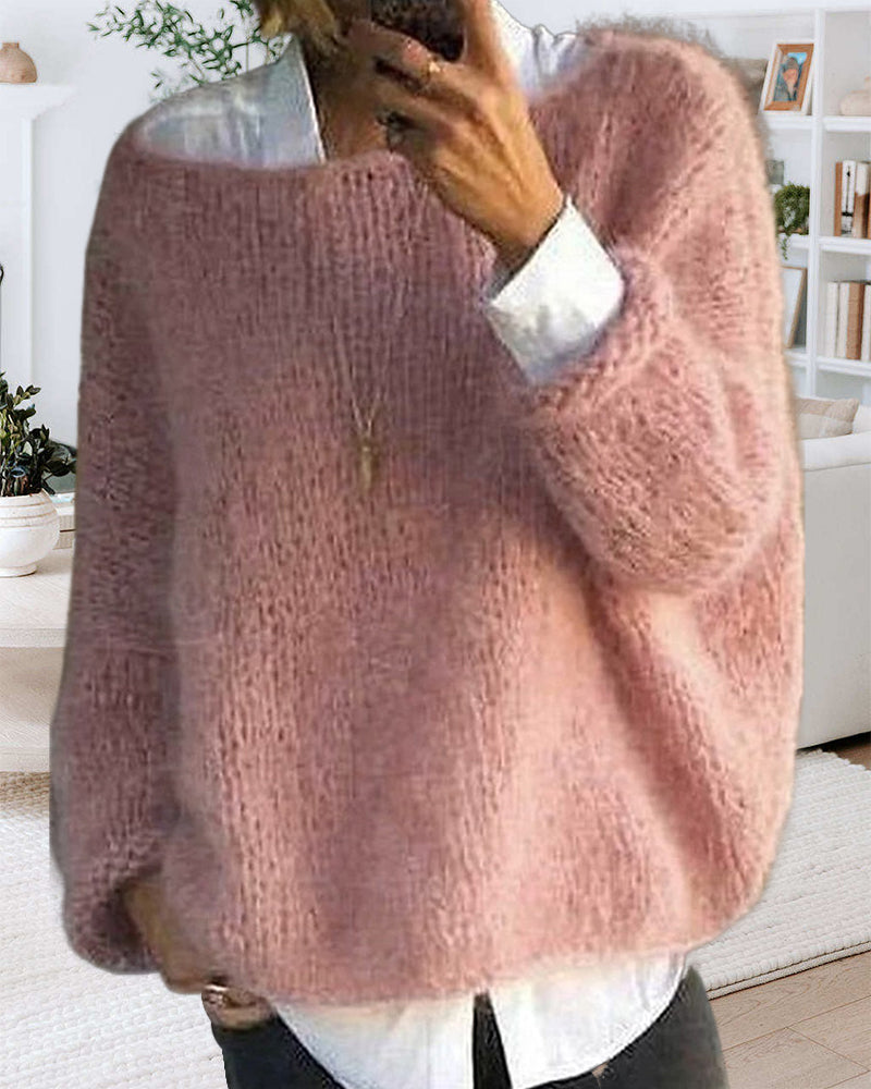 Femme portant un pull en laine rose doux, ample et texturé, sur une chemise blanche, dans un salon moderne. Mode automne-hiver, confort, style décontracté.