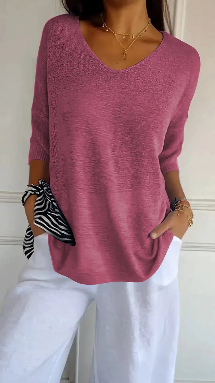 Femme portant un pull rose à col en V, manches trois-quarts, avec un pantalon blanc. Accessoire: foulard rayé noir et blanc au poignet.