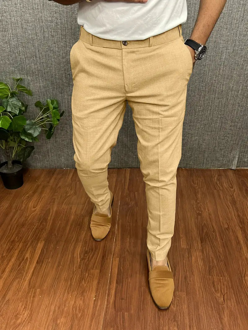 Pantalon beige élégant pour homme, porté avec des mocassins assortis et une montre noire, sur fond de sol en bois et plante verte.