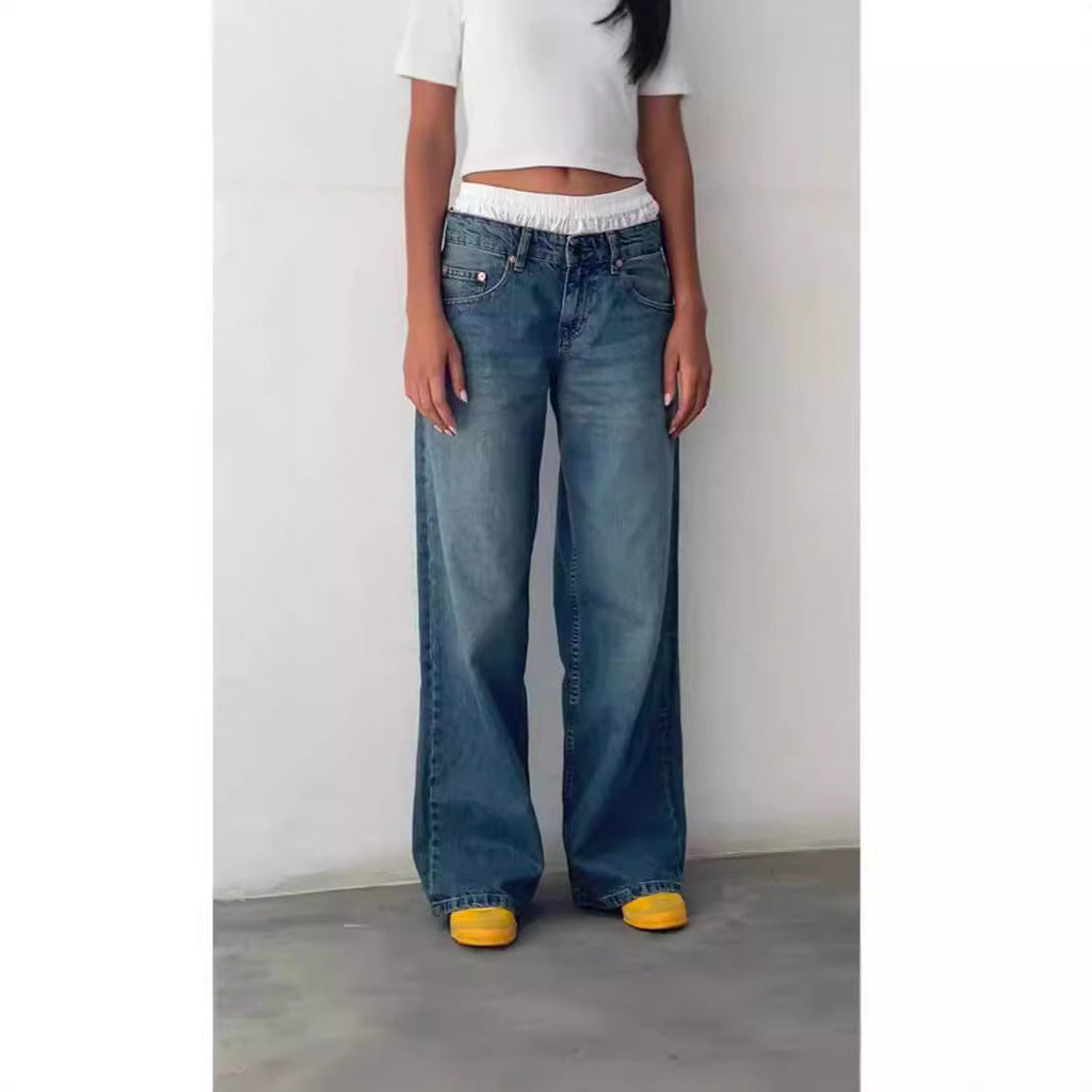 Femme portant un jean large bleu et un t-shirt blanc, debout contre un mur blanc, chaussures jaunes visibles. Mode décontractée, tendance streetwear.