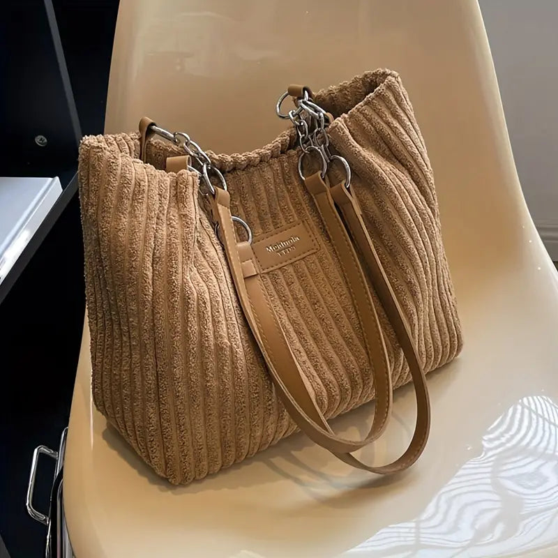 Sac à main en tissu côtelé beige sur une chaise blanche, avec anses en cuir et détails métalliques. Accessoire mode élégant et tendance.