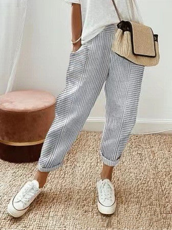 Femme portant un pantalon rayé bleu et blanc, avec un sac en paille et des baskets blanches, debout sur un tapis beige. Mode décontractée chic.