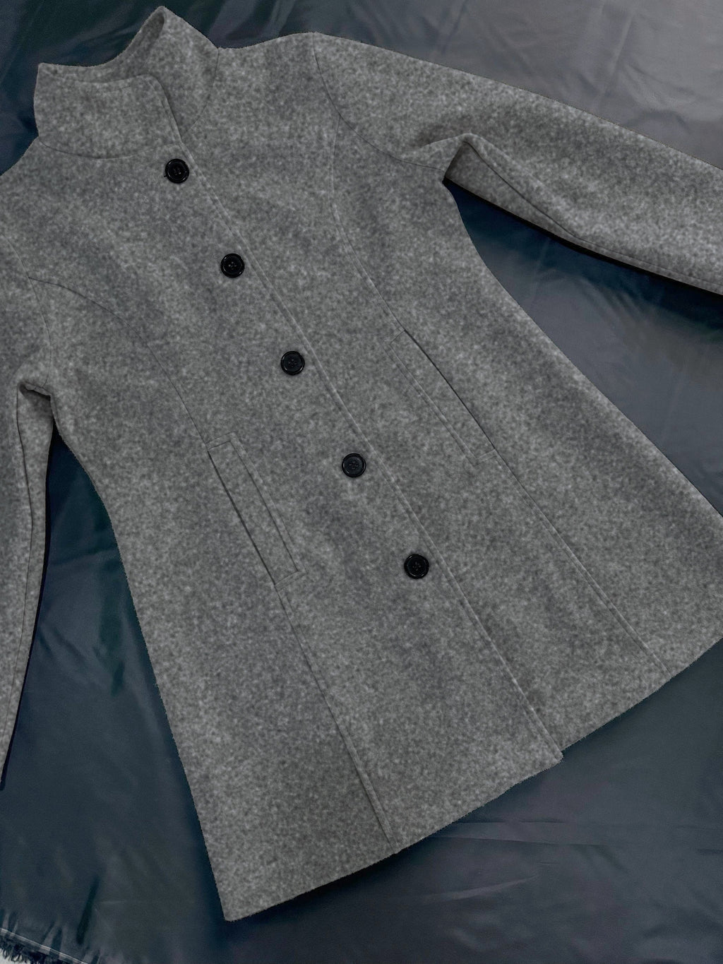 Manteau gris en laine pour femme, élégant et chaud, avec col montant et boutons noirs, posé sur une surface sombre. Mode hiver tendance.