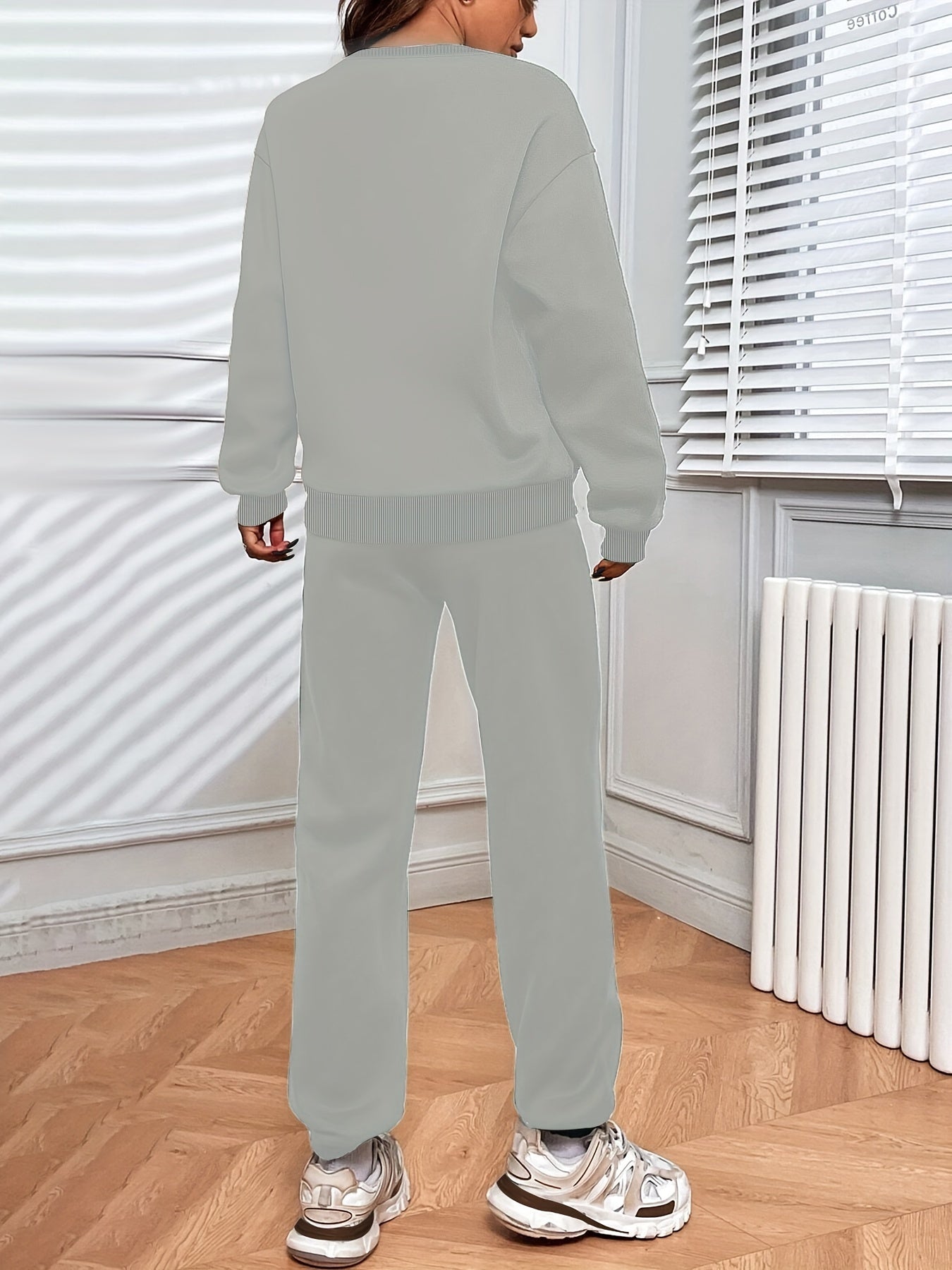 Personne portant un ensemble de survêtement gris clair, debout dans une pièce lumineuse avec parquet, radiateur blanc et stores vénitiens.