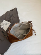 Sac à main en cuir marron ouvert sur une veste grise, avec un livre et des lunettes. Accessoire mode élégant, tendance et pratique.