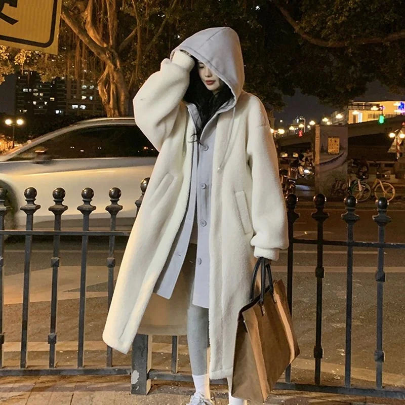 Femme portant un manteau long en laine blanc avec capuche, tenant un sac marron, debout près d'une rue éclairée la nuit. Mode hiver urbain.