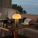 Lampe de table design champignon sur bureau moderne avec vue sur mer, livre ouvert, objets décoratifs en bois et métal, ambiance cosy et élégante.