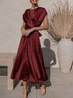 Robe longue en satin bordeaux, élégante et fluide, portée par une femme. Décor minimaliste avec vase blanc et plante séchée en arrière-plan.