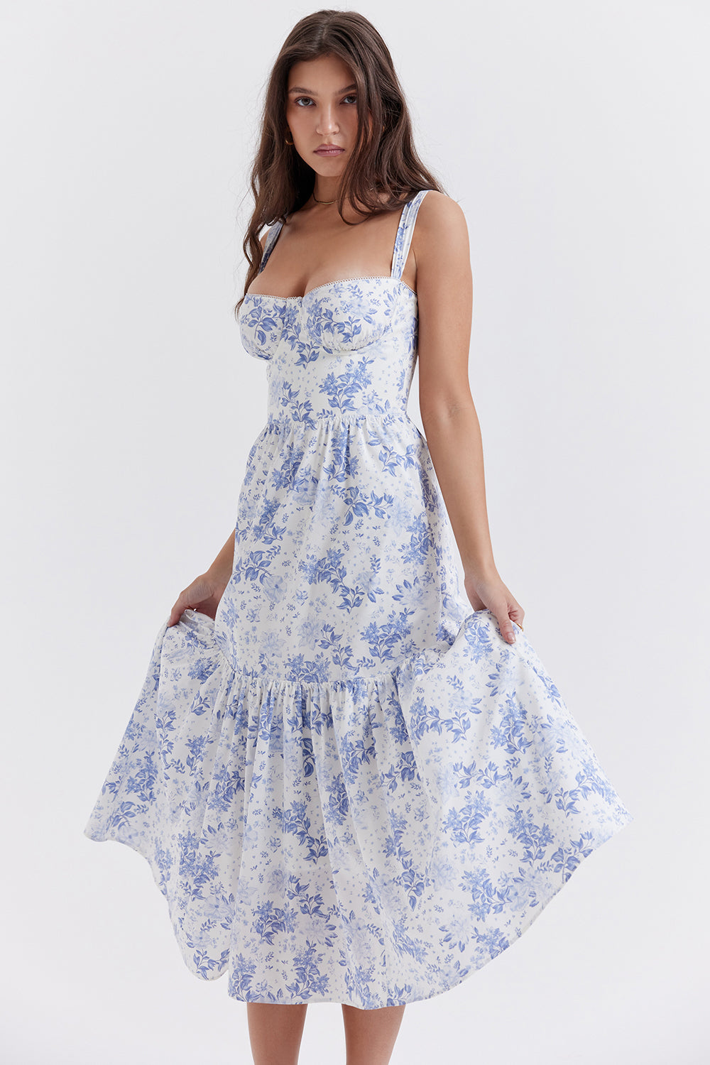 Robe longue à fleurs bleues et blanches portée par une femme, style été, bretelles fines, coupe évasée, fond blanc. Mode féminine élégante.