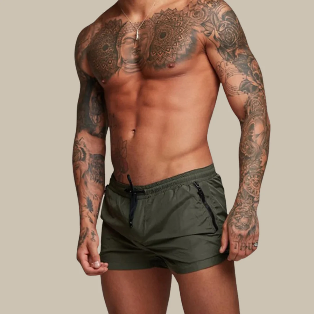Homme musclé torse nu avec tatouages détaillés, portant un short de bain vert kaki. Mode masculine, style tatouage, fitness.