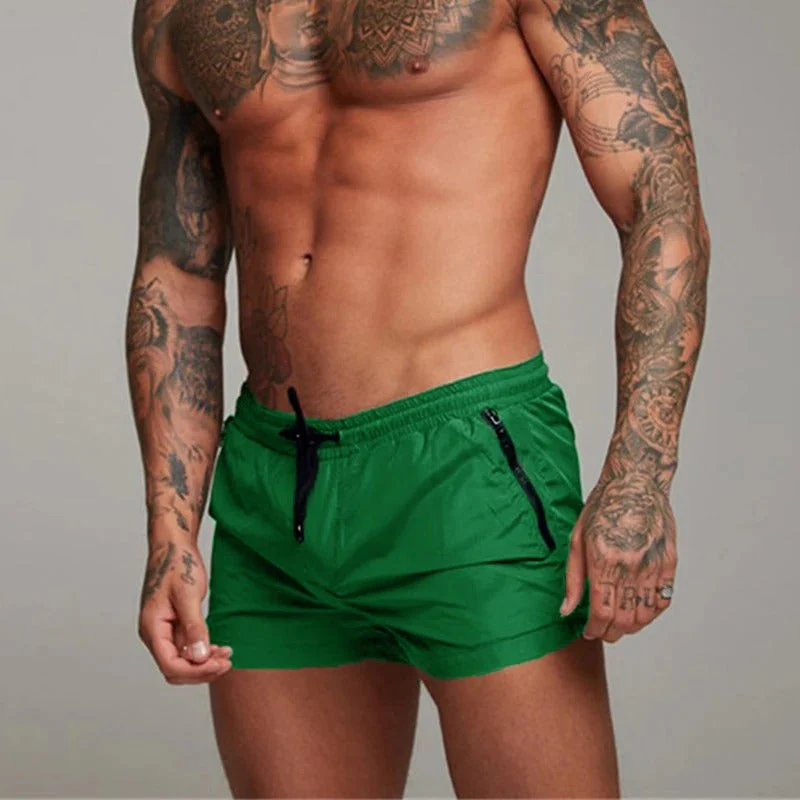 Homme musclé tatoué portant un short de bain vert, torse nu, posant sur fond neutre. Mode masculine, maillot de bain tendance, style estival.