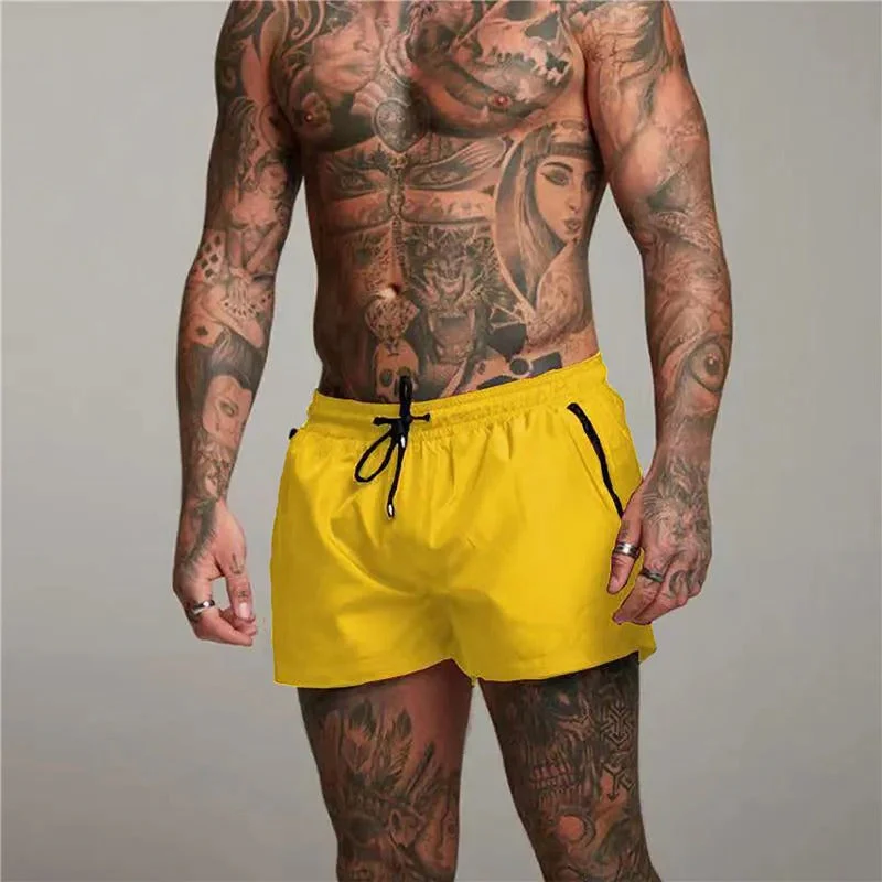 Homme musclé tatoué portant un short de bain jaune vif, posant debout. Mode plage, style estival, tenue de natation tendance.