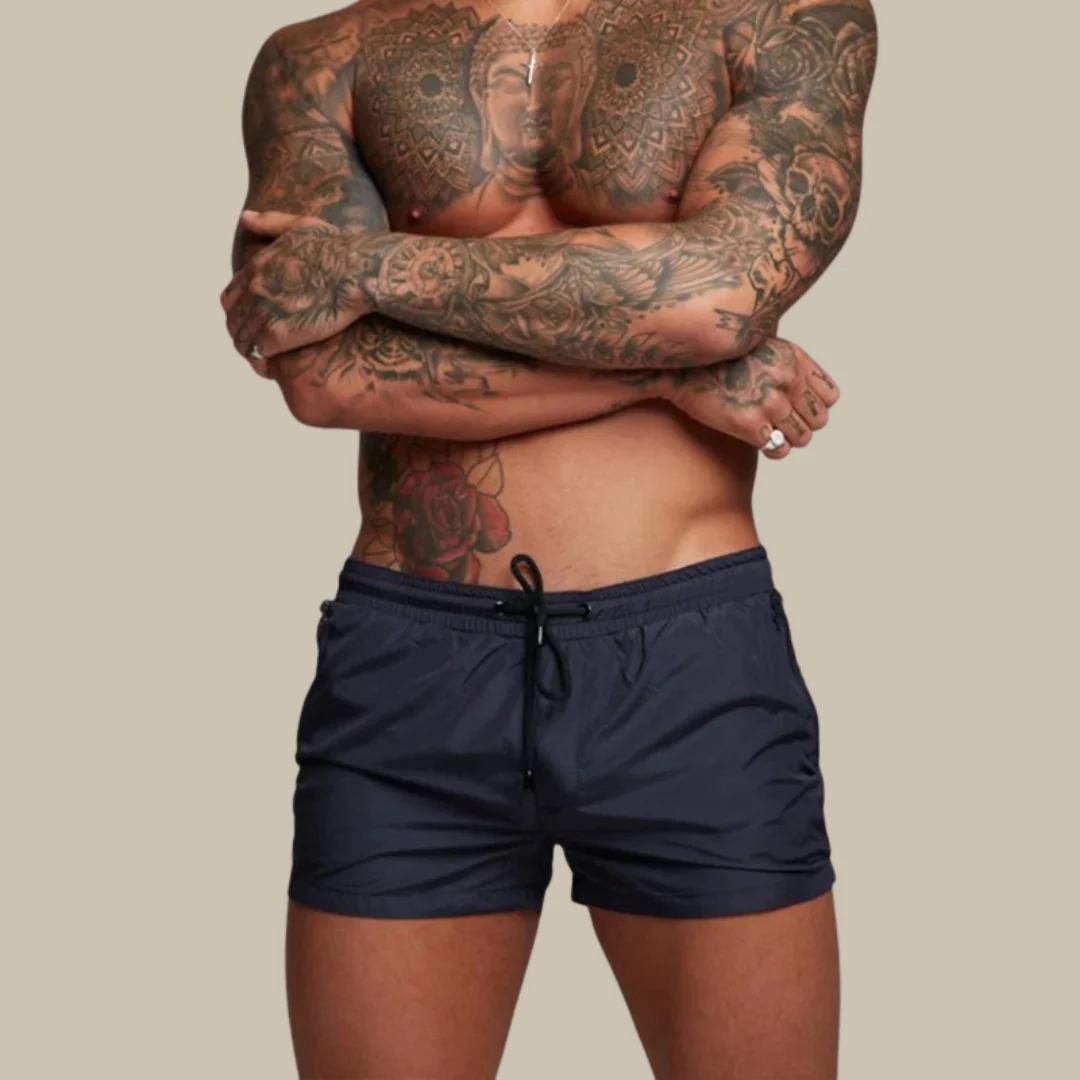 Homme torse nu avec tatouages détaillés, portant un short de bain bleu marine. Style moderne, mode masculine, tatouages artistiques.