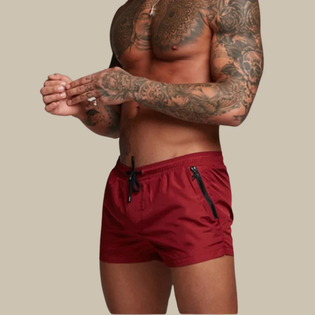 Homme torse nu tatoué portant un short de bain rouge avec cordon noir, fond beige. Mode masculine, style plage, tatouages.