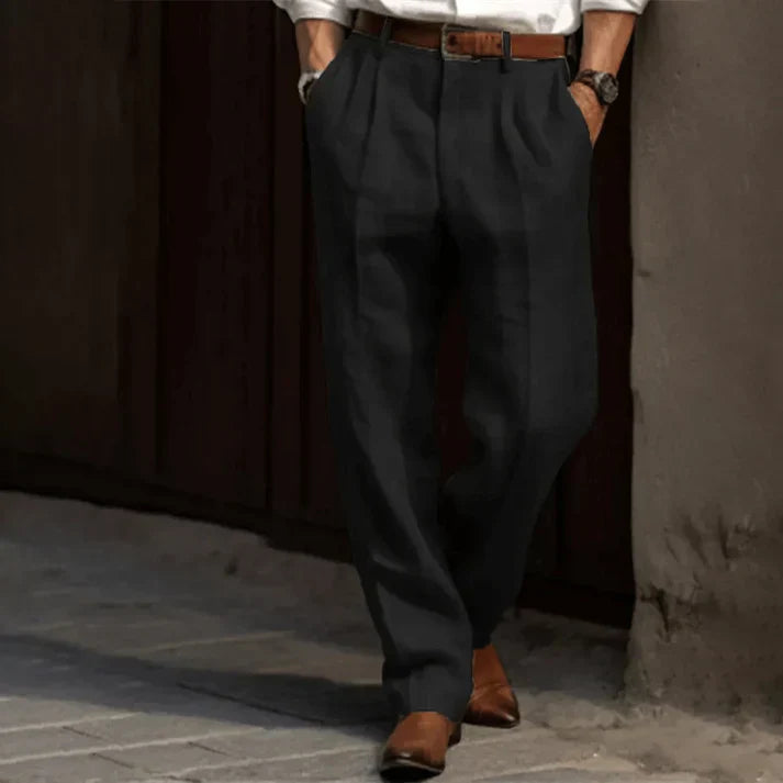 Homme en pantalon noir élégant, chemise blanche, ceinture marron, chaussures en cuir marron, appuyé contre un mur. Mode masculine chic et décontractée.