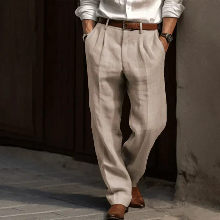Homme en pantalon beige en lin, chemise blanche, ceinture marron, chaussures en cuir marron, pose décontractée. Mode masculine élégante.