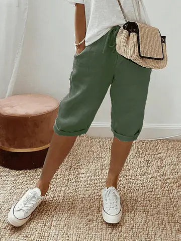 Femme portant un short vert kaki, t-shirt blanc, baskets blanches et sac à main en paille, posant sur un tapis beige. Mode décontractée estivale.