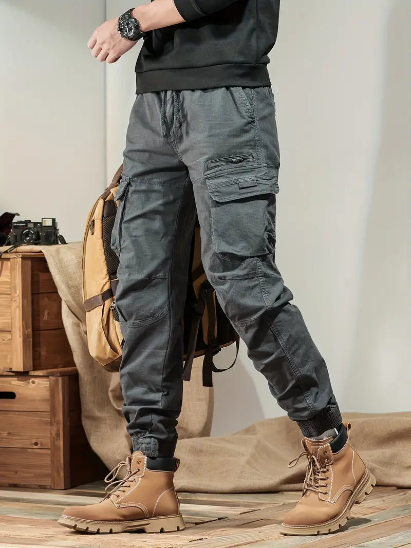 Homme portant un pantalon cargo gris, chaussures de randonnée marron, tenant un sac à dos beige. Mode outdoor, style décontracté, vêtements pratiques.