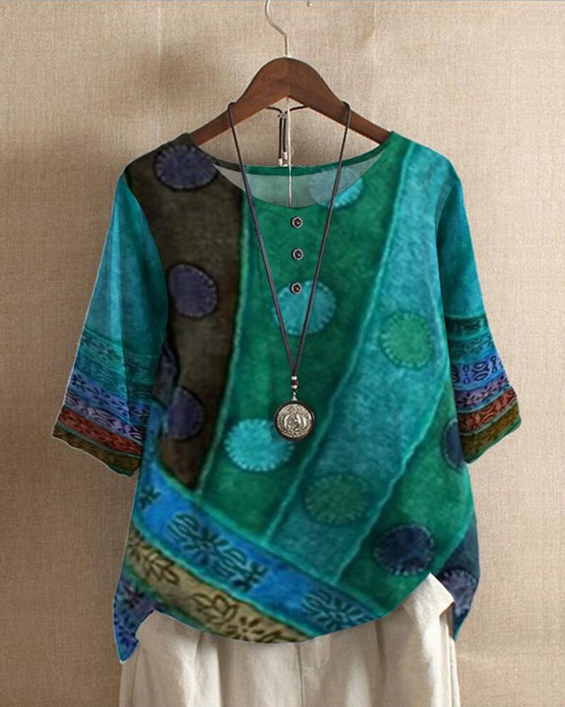 Blouse bohème chic turquoise à motifs ethniques, manches trois-quarts, col rond avec boutons, suspendue sur un cintre, collier pendentif argenté.