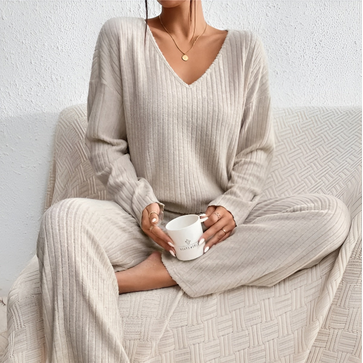 Femme en pyjama beige côtelé, assise sur un canapé, tenant une tasse blanche. Mode détente, confort à la maison, style cocooning élégant.