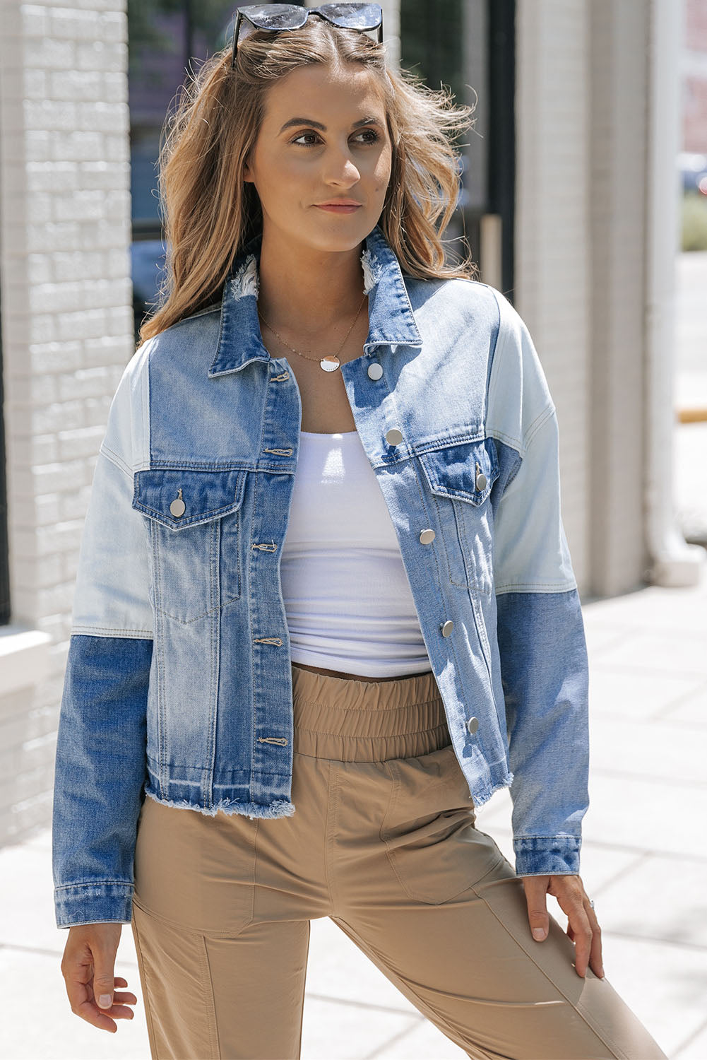 Femme portant une veste en jean patchwork, t-shirt blanc et pantalon beige, posant à l'extérieur par temps ensoleillé. Mode décontractée tendance.